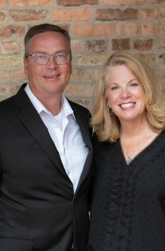 A man in a black suit and white shirt stands smiling next to a woman in a black blouse with blonde hair, also smiling. They are posing together in front of a brick wall. A man in a black suit and white shirt stands smiling next to a woman in a black blouse with blonde hair, also smiling. They are posing together in front of a brick wall.