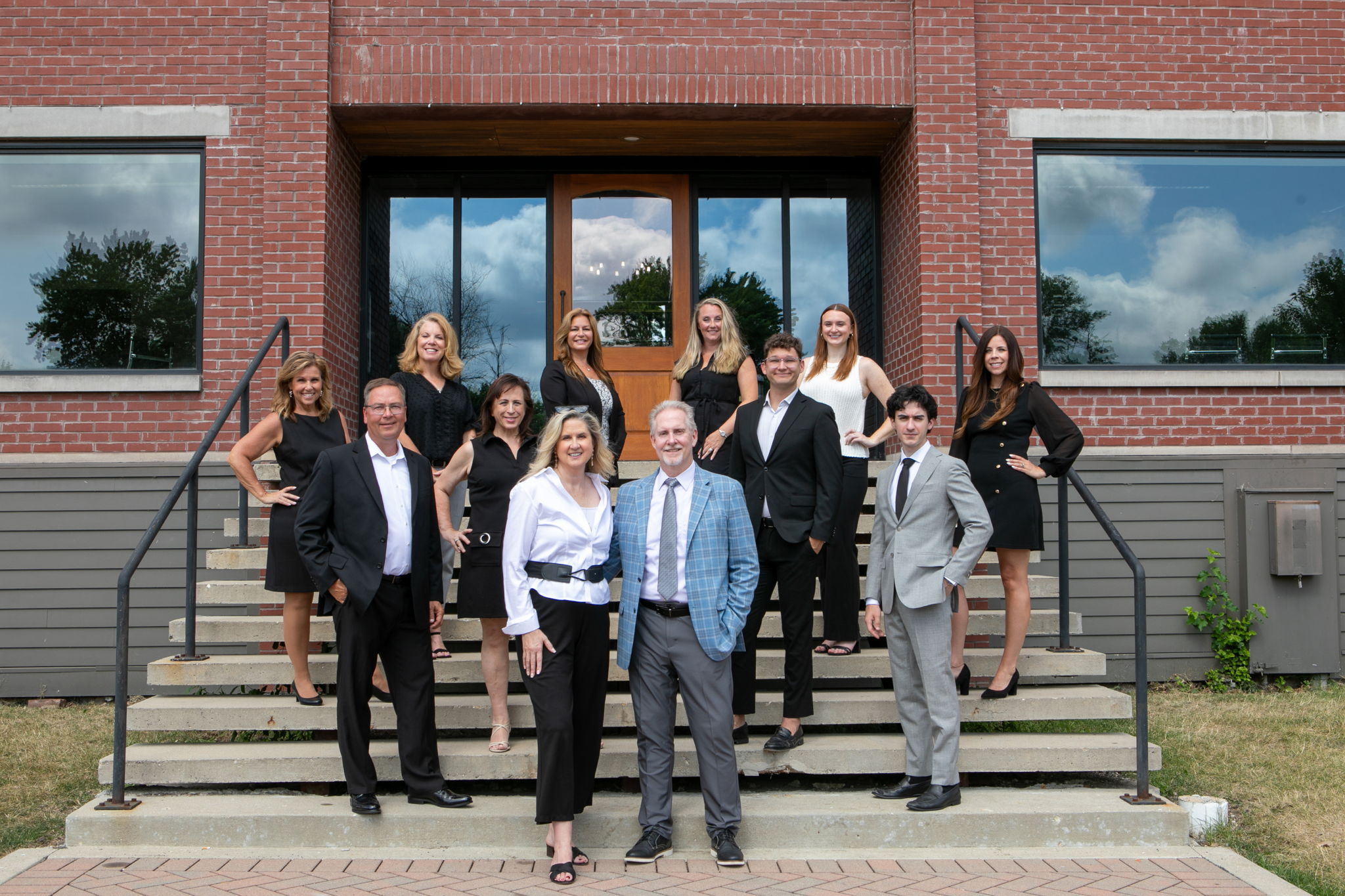 A group of twelve professionally dressed people pose and smile on the steps outside a brick building with large windows and a wooden door.