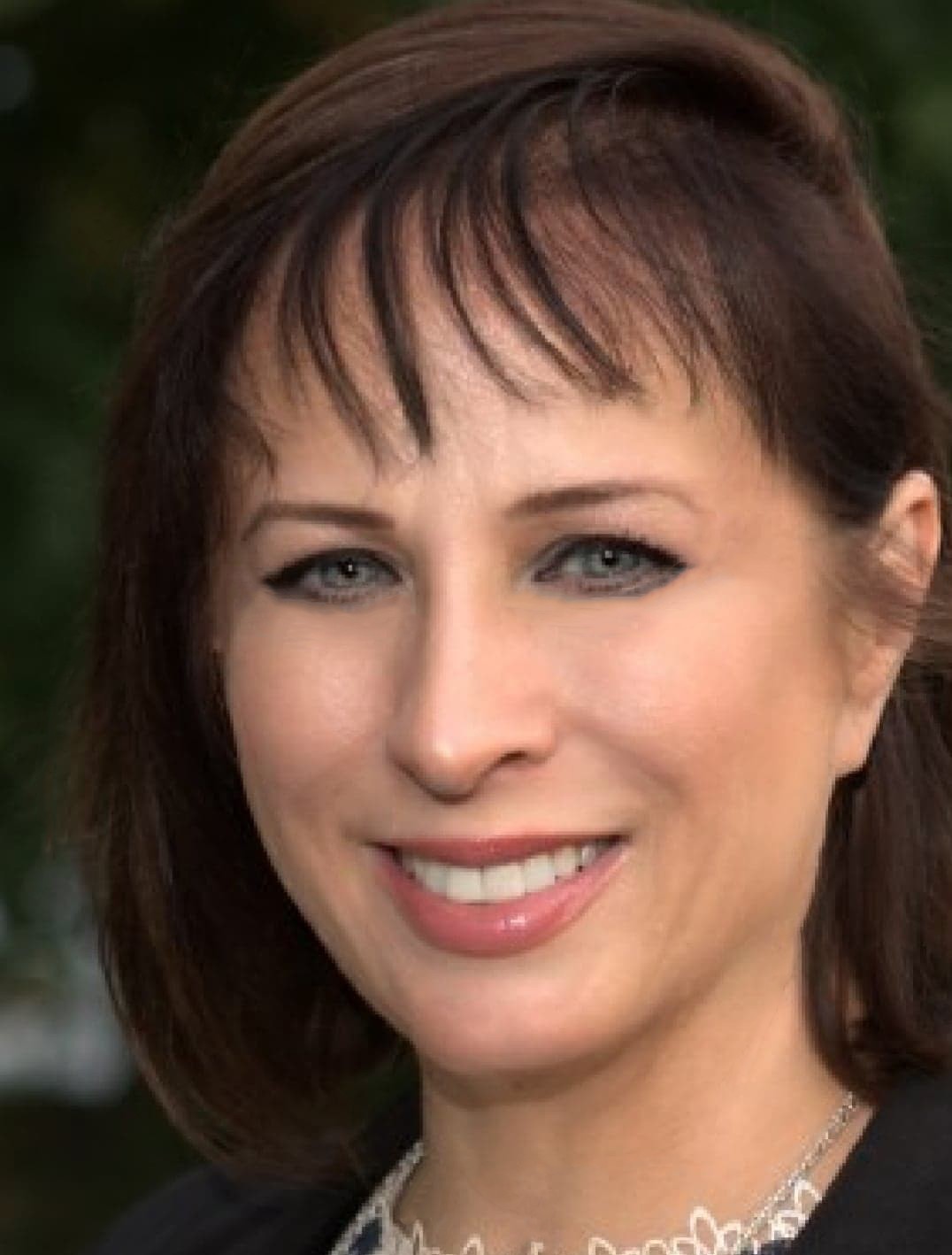 A woman with straight brown hair and bangs is smiling outdoors. She has light skin, blue eyes with black eyeliner, and is wearing a lacy white top and a dark jacket. Green foliage is blurred in the background. A woman with straight brown hair and bangs is smiling outdoors. She has light skin, blue eyes with black eyeliner, and is wearing a lacy white top and a dark jacket. Green foliage is blurred in the background.